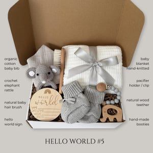 Puede incluir: Un set de regalo para beb&eacute; en una caja de cart&oacute;n que contiene una manta blanca de punto gofrado con una cinta gris, un sonajero de elefante de ganchillo gris, un par de patucos de punto grises, un cepillo de madera, un clip de silicona gris para chupete, un mordedor de elefante de madera y un disco de madera con el texto Hello World.