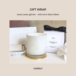 May include: A white candle in a glass jar with a gold base, sitting on a white surface. The candle is labeled "CandleCard by CANDLY". The candle is packaged in a white box with a black ribbon.