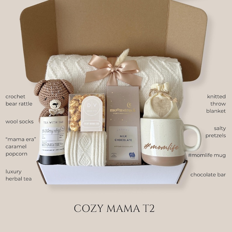 May include: A gift box with a cream-colored knitted throw blanket, a crochet bear rattle, a mug with "#momlife" text, a chocolate bar, and other items. The box includes wool socks, caramel popcorn, and herbal tea.