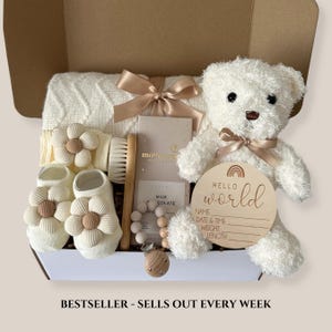 May include: A baby gift box filled with a white teddy bear, a wooden sign that says "Hello World", baby booties, a blanket, a brush, a pacifier, and a chocolate bar. The box is beige and has a beige bow.