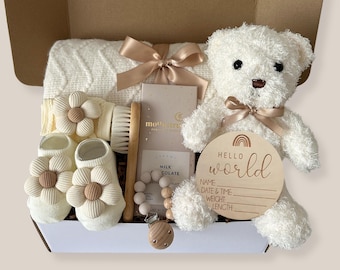 Puede incluir: Una caja de regalo que contiene una manta de punto color crema con cinta color champán, un oso de peluche blanco con lazo a juego, una placa de madera Hello World, una barra de chocolate con leche, un cepillo de madera y patucos color crema.
