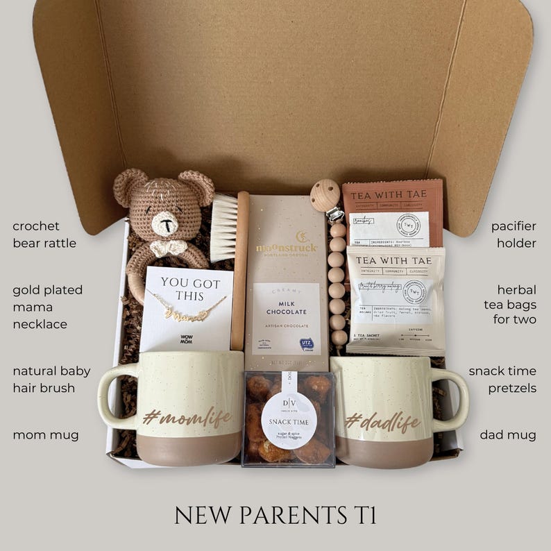May include: A gift box for new parents, containing a crochet bear rattle, gold-plated mama necklace, natural baby hair brush, two mugs with the hashtags #momlife and #dadlife, milk chocolate, herbal tea bags, snack pretzels, and a pacifier holder.