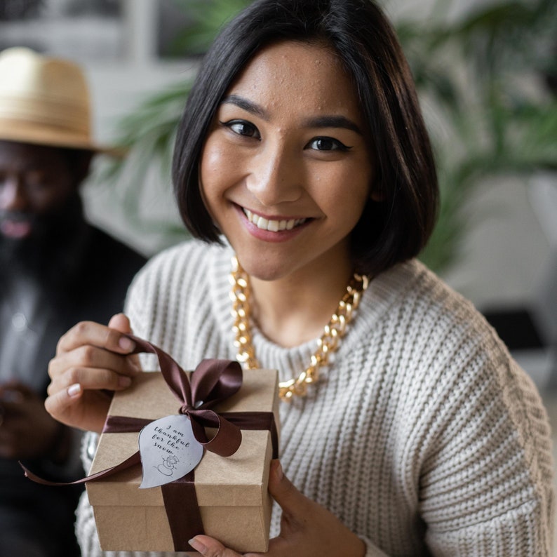 May include: A woman is holding a gift box wrapped in brown paper with a brown ribbon and a white tag that says "I am thankful for the small things".