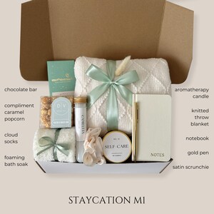May include: A gift box filled with self-care items. Includes a white knitted throw blanket with a mint green ribbon, a notebook, a gold pen, a candle, and other items. The box is labeled "STAYCATION MI".