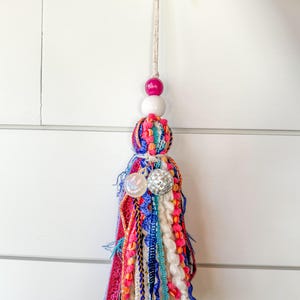 May include: A colorful tassel with a white, pink, and green bead. The tassel is made of various fabrics, including yarn, ribbon, and fringe. It is hanging from a white wall.