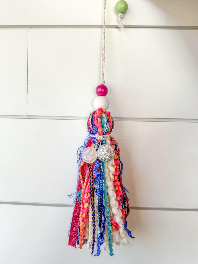 May include: A colorful tassel with a white, pink, and green bead. The tassel is made of various fabrics, including yarn, ribbon, and fringe. It is hanging from a white wall.