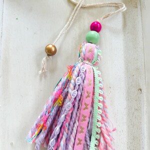 May include: A pink and purple tassel with a gold, green, and pink bead accent. The tassel is made of yarn and has a variety of textures and colors.