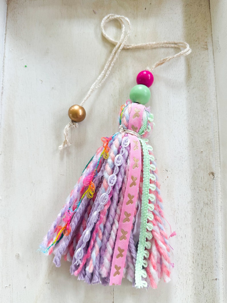 May include: A pink and purple tassel with a gold, green, and pink bead accent. The tassel is made of yarn and has a variety of textures and colors.