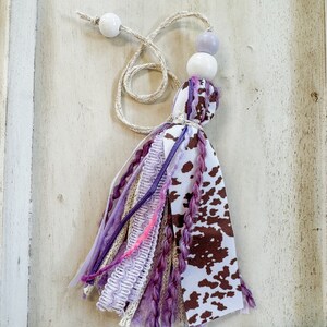 May include: A purple and brown tassel with a cow print ribbon and white beads. The tassel is made of various fabrics and yarns.