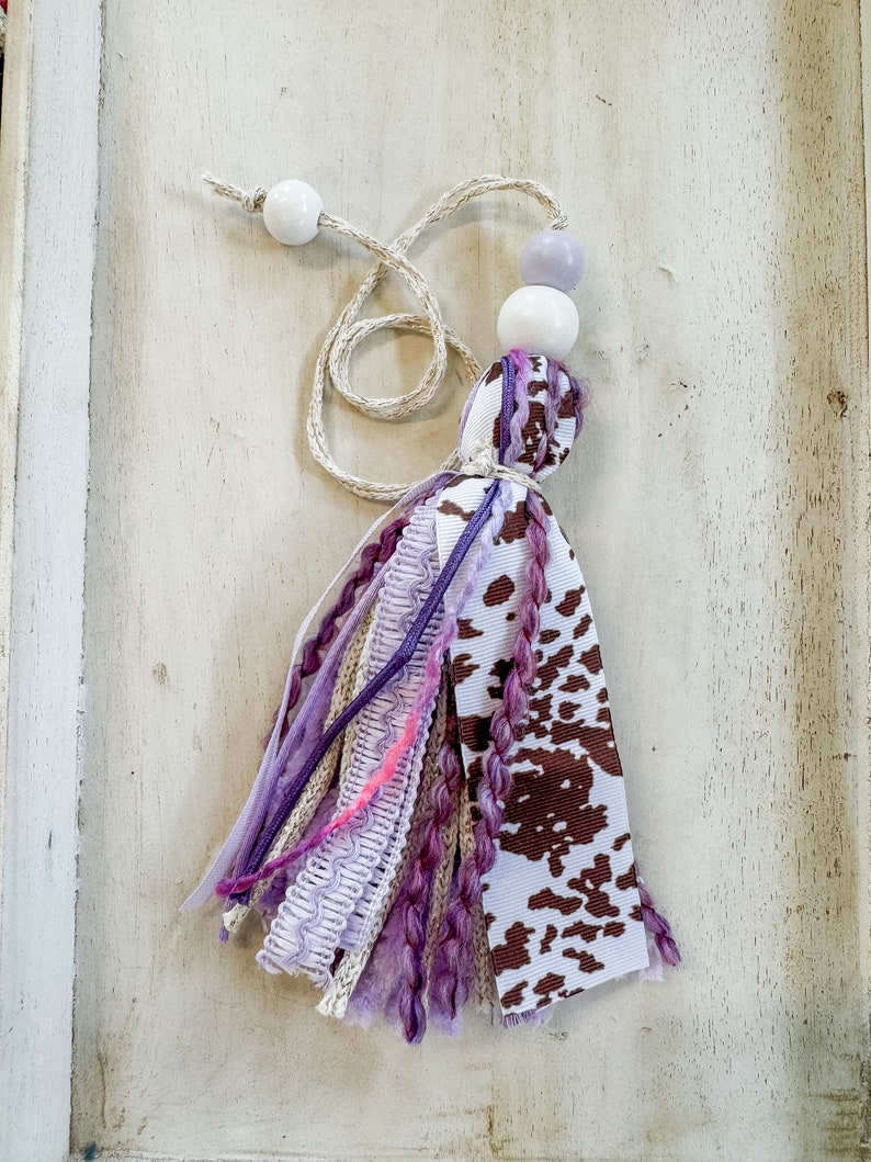 May include: A purple and brown tassel with a cow print ribbon and white beads. The tassel is made of various fabrics and yarns.