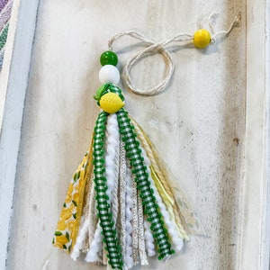 May include: A green, white, and yellow tassel with a lemon charm. The tassel is made of various ribbons, yarn, and fabric. It is hanging from a white string with a yellow bead at the end.