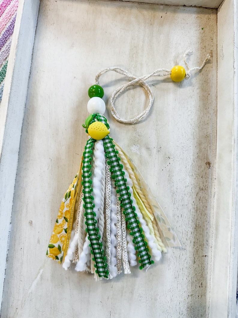 May include: A green, white, and yellow tassel with a lemon charm. The tassel is made of various ribbons, yarn, and fabric. It is hanging from a white string with a yellow bead at the end.