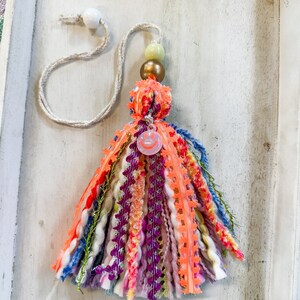 May include: A colorful tassel keychain with a pink smiley face charm, green and gold beads, and a white cord.