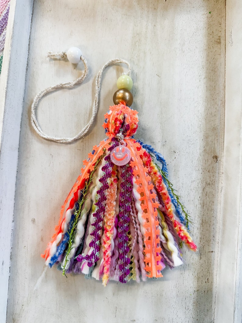 May include: A colorful tassel keychain with a pink smiley face charm, green and gold beads, and a white cord.