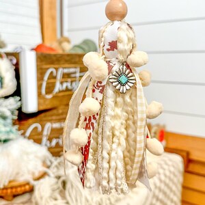 May include: A white and red tassel ornament with a turquoise and silver charm. The tassel is made of yarn, ribbon, and pom-poms. It is hanging from a wooden bead and gold bead.
