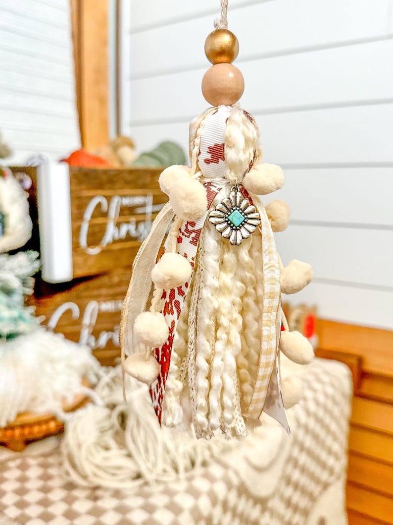 May include: A white and red tassel ornament with a turquoise and silver charm. The tassel is made of yarn, ribbon, and pom-poms. It is hanging from a wooden bead and gold bead.