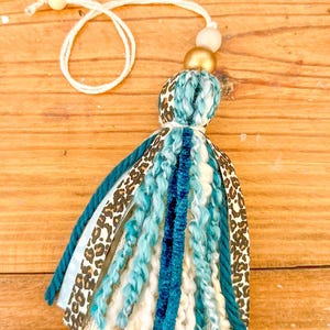 May include: A teal, white, and brown tassel with a leopard print ribbon and a gold and beige bead at the top. The tassel is hanging from a white string.