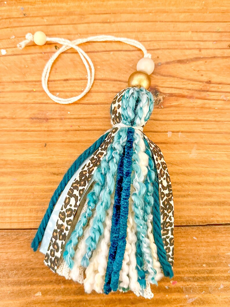 May include: A teal, white, and brown tassel with a leopard print ribbon and a gold and beige bead at the top. The tassel is hanging from a white string.