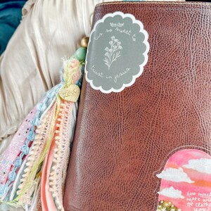May include: A brown leather-bound journal with a gray sticker that says "Tis so sweet to trust in Jesus" and a floral design. The journal has a colorful tassel attached to it.