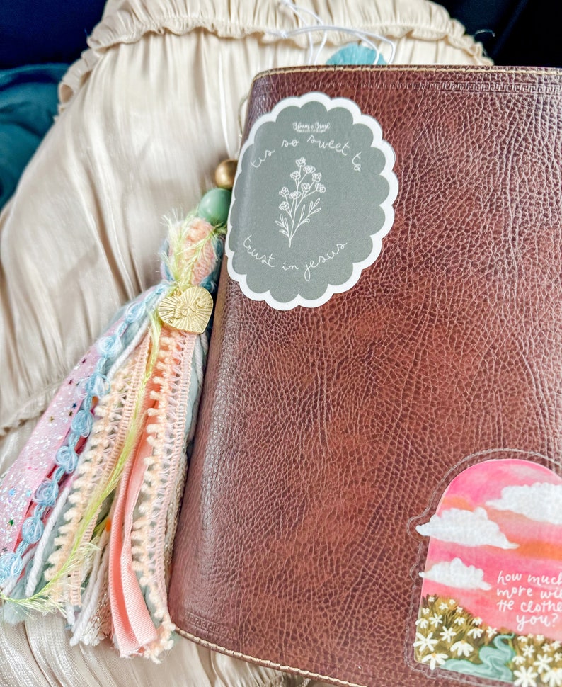 May include: A brown leather-bound journal with a gray sticker that says "Tis so sweet to trust in Jesus" and a floral design. The journal has a colorful tassel attached to it.