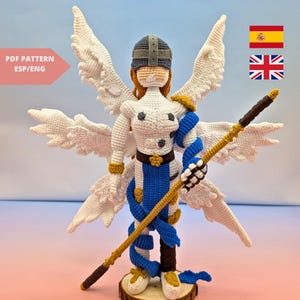 Puede incluir: Figura de ángel tejida a ganchillo con grandes alas blancas, que lleva un casco y una armadura, y sostiene un bastón. La figura es principalmente blanca con detalles azules y dorados. El texto "PDF PATTERN ESP/ENG" es visible.
