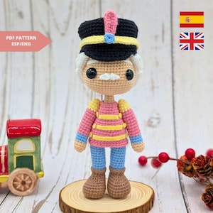 May include: A handmade crochet doll of a nutcracker soldier. The doll wears a black hat with yellow trim, a pink jacket with yellow stripes, blue pants, and brown boots. The text "PDF PATTERN ESP/ENG" is in the upper left corner.