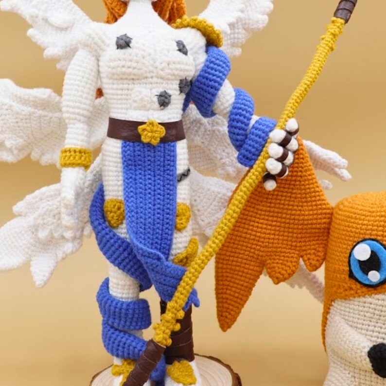 以下が含まれることがあります： 白い翼、青と白の衣装、黄色の杖を持つキャラクターの編みぐるみ。フィギュアは、黄色の花の形をしたバックルが付いた茶色のベルトをしています。フィギュアは木の台座の上に立っています。別のオレンジと白の編みぐるみのキャラクターが部分的に見えます。