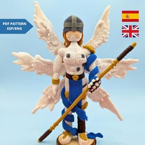 Puede incluir: Una figura de ángel de ganchillo con grandes alas blancas, un atuendo azul y blanco y un bastón dorado. El ángel tiene un casco y pelo rojo. La figura se exhibe sobre una base de madera. La imagen también incluye el texto "PDF Pattern ESP/ENG".