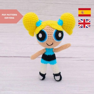 Puede incluir: Muñeca de crochet de un personaje de dibujos animados con cabello rubio, ojos azules y un vestido a rayas azul y negro. La muñeca está de pie sobre una superficie de madera blanca.