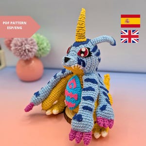 May include: A crocheted blue and yellow dinosaur with a pink and blue chest plate. The dinosaur has a yellow horn and red eyes.
