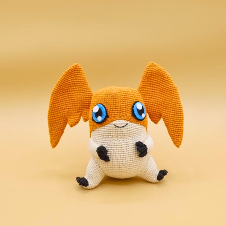 以下が含まれることがあります： オレンジ、白、黒色の手作りの編みぐるみデジモンぬいぐるみ。 ぬいぐるみは大きなオレンジ色の翼、青い目、黒い足を持っています。 デジモンは無地の黄色の背景に座っています。