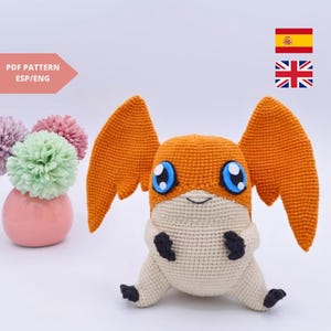 May include: A handmade orange and cream-colored crochet creature with large wings and blue eyes. The plush toy has black accents on its feet and arms. The image also includes a vase of flowers and a text that says "PDF PATTERN ESP/ENG".