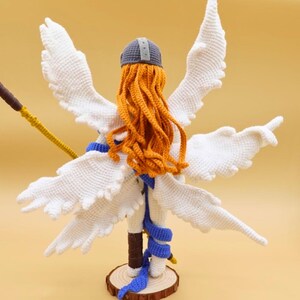 以下が含まれることがあります： 白い翼、オレンジ色の髪、灰色のヘルメットを持つ手作りのかぎ針編みの天使人形。 人形は青いサッシュを着ており、黄色と茶色の杖を持っています。 木製の台座に取り付けられています。