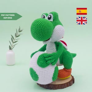 Puede incluir: Muñeco de Yoshi tejido a ganchillo en verde, blanco, rojo y naranja. El muñeco sostiene un gran huevo blanco y verde. Está sobre una pequeña base de madera. La imagen incluye el texto "PDF PATTERN ESP/ENG".