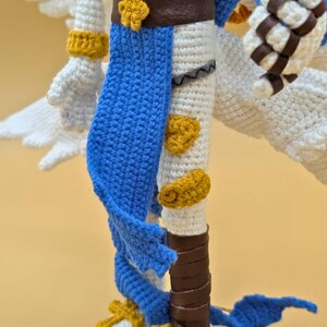 以下が含まれることがあります： 白、青、金色のアクセントが施された手作りのかぎ針編みフィギュア。白いボディに青と金のディテール、茶色の革のアクセントがあります。翼は白で、手作りの工芸品です。