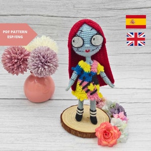 Puede incluir: Muñeca de crochet con pelo rojo, un vestido de patchwork azul, amarillo y rosa, y calcetines a rayas blancas y negras. La muñeca está de pie sobre una base redonda de madera con flores alrededor.