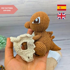Puede incluir: Un juguete de dinosaurio marrón tejido a ganchillo con una máscara de calavera color crema. El dinosaurio tiene un diseño detallado, incluyendo una cola y espinas. El texto "PDF PATTERN ESP/ENG" es visible en la esquina superior izquierda.