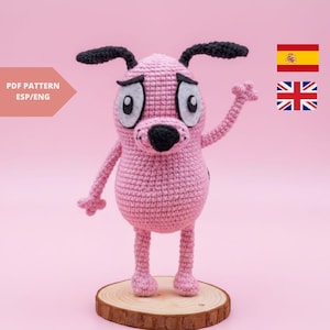 May include: A pink crocheted Courage the Cowardly Dog figure with black ears and nose, and large, sad eyes. The figure is standing on a wooden disc against a pink background. The text "PDF PATTERN ESP/ENG" is on the left.
