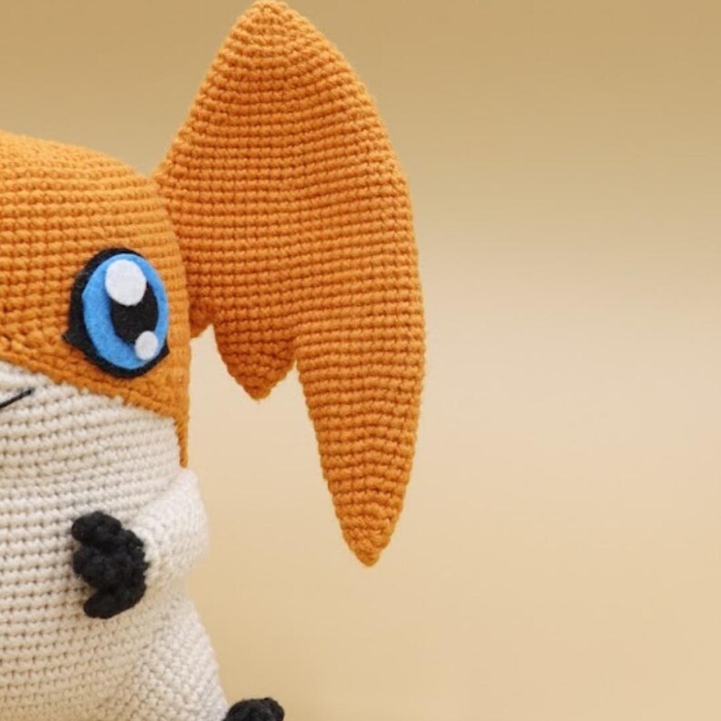 以下が含まれることがあります： オレンジ色の耳、白い顔、青い目をしたキャラクターを特徴とする、かぎ針編みのぬいぐるみ。おもちゃは糸でできており、質感のある外観をしています。デザインは漫画のキャラクターを彷彿とさせます。