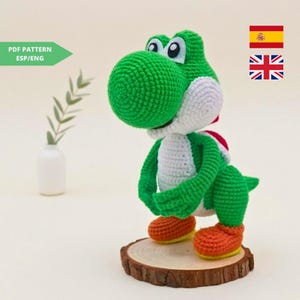 Puede incluir: Figura de Yoshi hecha a mano con ganchillo, con cuerpo verde, vientre blanco y silla de montar roja. La figura está sobre una base de madera con pies naranjas y amarillos. Incluye patrón PDF en inglés y español.