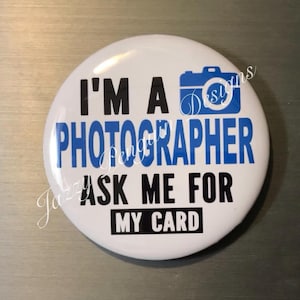 May include: A white button with the text "I'M A PHOTOGRAPHER ASK ME FOR MY CARD" in black and blue. A blue camera icon is at the top. The button is round and glossy.