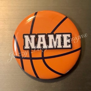 May include: Round orange button with a basketball design. The button features black lines mimicking a basketball and the word "NAME" in white block letters with a black border. The button has the text "Jazzy Lady Designs" in cursive.
