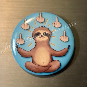 May include: A round, light blue button featuring a cartoon sloth in a meditative pose, surrounded by hands giving the middle finger. The sloth is brown with a light brown face. The button has the text "Jazzy Penguin Designs".