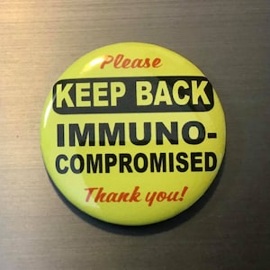 May include: A round, yellow button with black text that reads "KEEP BACK IMMUNO-COMPROMISED." The words "Please" and "Thank you!" are in red. The button is likely a safety or awareness item.