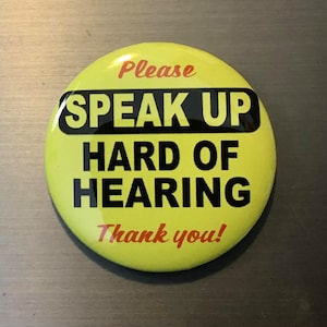 May include: A round, yellow button with the words "PLEASE SPEAK UP HARD OF HEARING Thank you!" in black and red text. The button has a glossy finish and is likely a pin or badge.