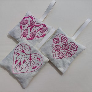May include: Three white fabric heart-shaped sachets with pink embroidered floral designs. The sachets have white ribbon loops for hanging.
