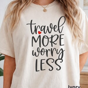 May include: An ivory short-sleeved t-shirt featuring the black text "travel MORE worry LESS" with a small red heart icon positioned between the words travel and MORE. The shirt has a crew neckline and a relaxed fit, showcasing a simple design.