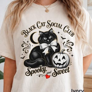 May include: An ivory t-shirt featuring a graphic of a black cat wearing a white bow, a crescent moon, a jack-o'-lantern, and bats. The text reads "Black Cat Social Club" and "Spooky and Sweet" with a small red heart icon below the text.