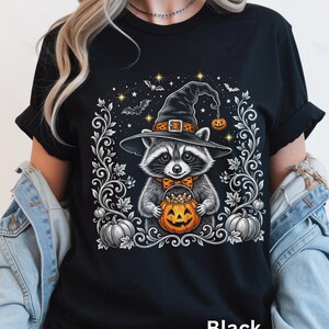 May include: A black t-shirt featuring a graphic of a raccoon wearing a witch hat and an orange bow tie. The raccoon holds a jack-o'-lantern bucket filled with candy corn. The design includes silver filigree, bats, and stars on a black background.