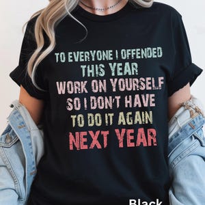 Op de afbeelding: Een zwart t-shirt met korte mouwen en de tekst "TO EVERYONE I OFFENDED THIS YEAR WORK ON YOURSELF SO I DON'T HAVE TO DO IT AGAIN NEXT YEAR" gedrukt in een verweerd, veelkleurig lettertype in lichtgroen, roze en rood.